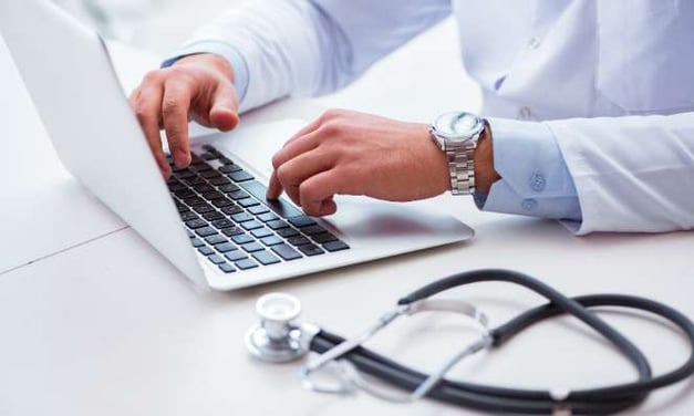 Person seated at a desk, with hands on a laptop keyboard, wearing a watch. A stethoscope is in the foreground on the desk.