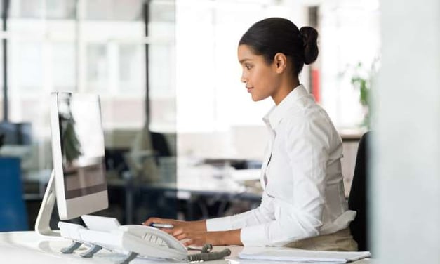 Woman with hair in a bun, wearing long sleeve white shirt, looking at the laptop monitor that's on the desk she's seated at. Windows in background.