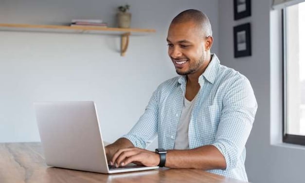 Man smiling at the laptop he's typing on. He is seated at a wooden desk or table. There is a single wooden shelf on the white wall in the background.