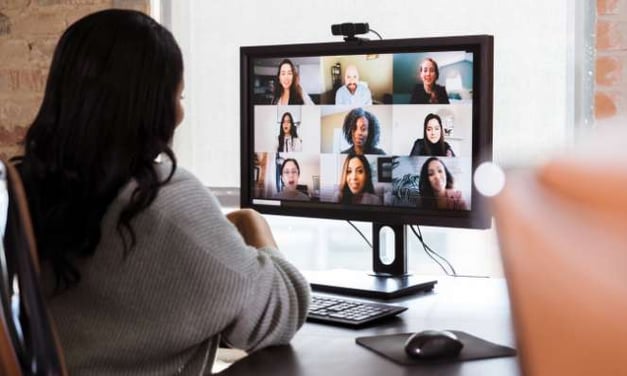Woman seated at desk looking at computer monitor for video call with many faces on the screen.