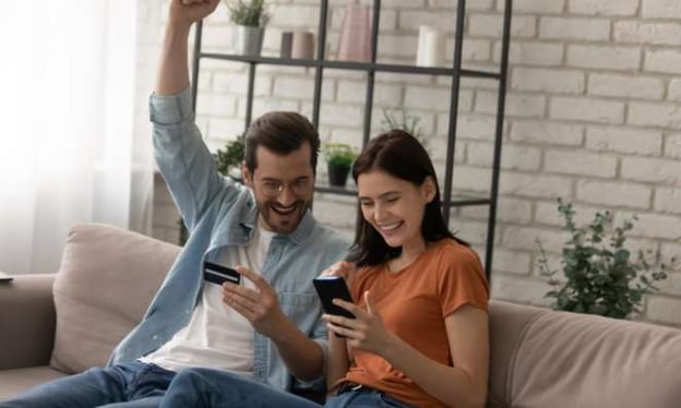 A man and woman are seated on a pink couch, looking at a phone and smiling. There is a brick wall, potted plant, and shelving in the background.