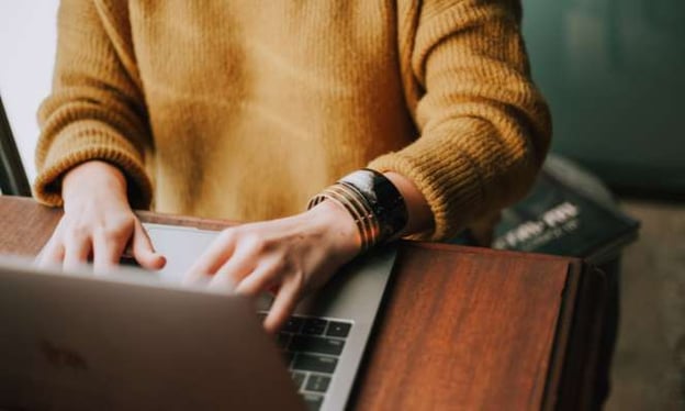 A woman in an orange sweater is sitting art a desk working on a laptop. 