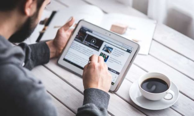 A man sits at a table while scrolling on his tablet, looking at articles, texts, and videos. Beside him is a cup of coffee.