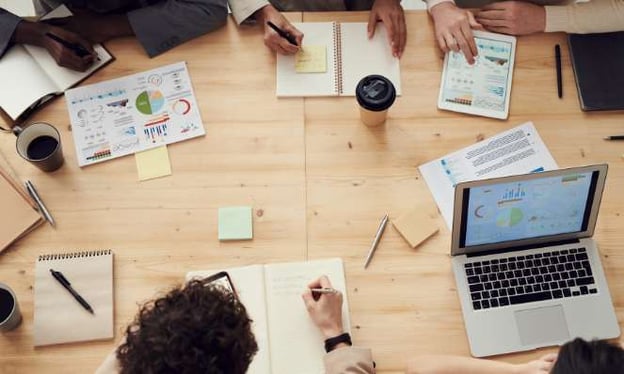 Five people are sitting around a table working. On the table is a data sheet, a notebook, a tablet, a laptop, a drink, sticky notepads, and a coffee. 