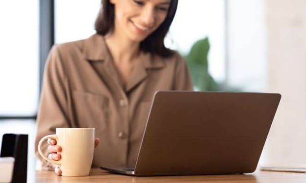 A woman is sitting at a desk working on her laptop while smiling. In her right hand is a coffee mug. She is wearing a brown blouse. 