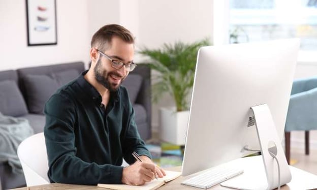 A man sits at a desk wearing glasses and smiles as he writes notes in a notebook. On the desk is a monitor and keyboard. 