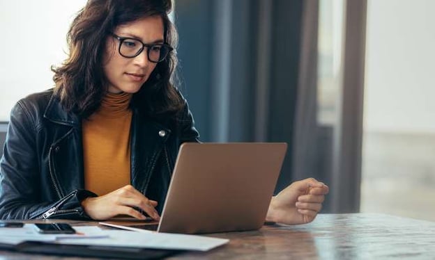 A woman is sitting at a table working on a laptop. She is wearing glasses, an orange top, and a jacket. There is a stack of pages on the table. 