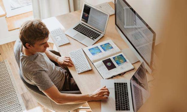 A man sits at a desk with three monitors in front of him, two are laptops. There is a keyboard and an open notebook. He is wearing a gray shirt.