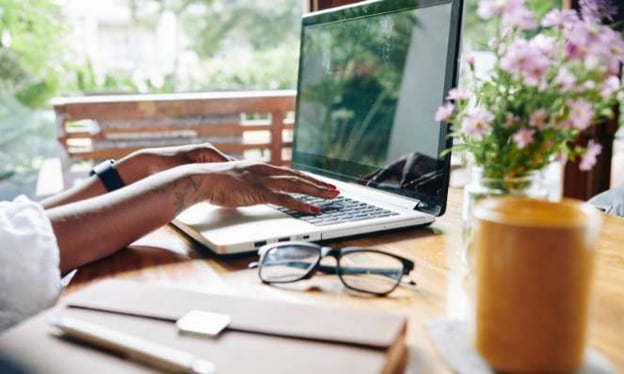 A woman's hands are typing on the keyboard of an open laptop while sitting at a desk. On the desk are glasses, a plant, and a book. 