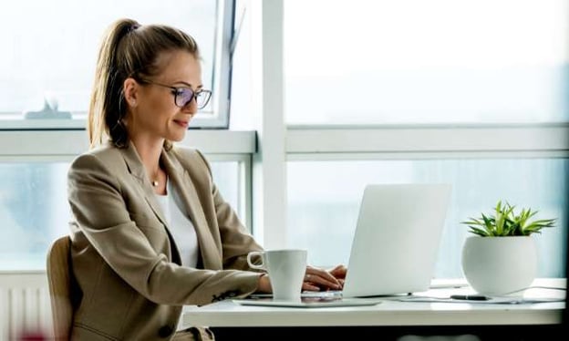 A woman wearing glasses sits at a desk, working on a laptop. A tablet, coffee cup, and plant are on the desk. There are windows behind her.