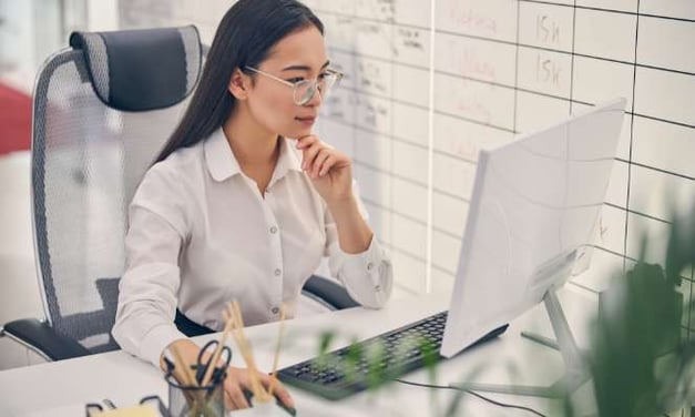 A woman sitting at a desk looking at a computer monitor while wearing glasses and looking puzzled. There is a container of pens on the desk.