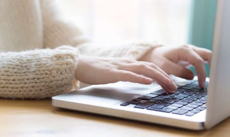 Hands typing on a laptop that is on a wooden desk or table. The person is wearing a cable knit sweater. There is a window in the background.