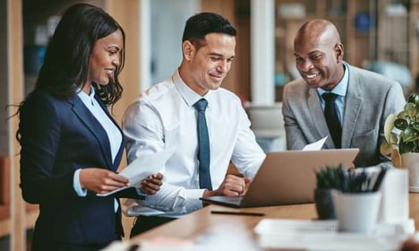 Three people in business attire standing around a table, looking at a laptop. Two of them are also holding paperwork. All are smiling.