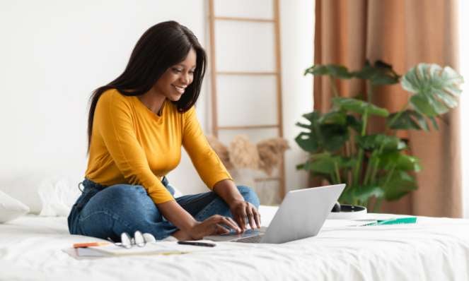 A woman sits on a bed, wearing jeans and a yellow top, and is looking at and typing on a laptop while smiling. A plant is in the background.