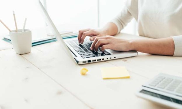 A man's hands are typing on a laptop keyboard, seated at a desk. A cup holding pens, a sticky notepad, and a calculator are on the desk. 