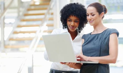 Two woman standing, one is holding a laptop, they are both looking at the screen. There are stairs in the background of the image.