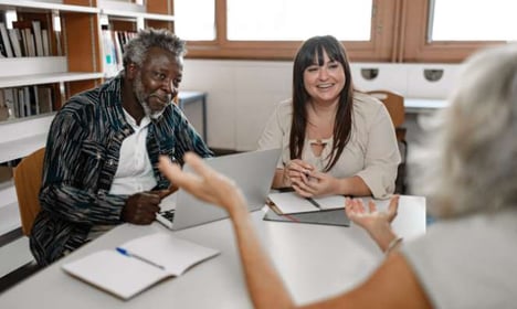 Three people seated at a table, two are facing the other in the foreground. There is an open laptop on the table and the people are smiling.