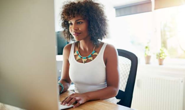 A woman sitting at a desk, looking at a computer monitor. She has curly hair and is wearing a necklace. A window and potted plants in background.