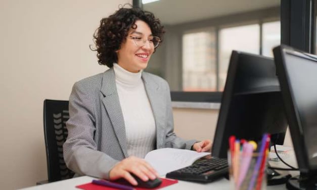 Woman with curly hair and glasses is smiling while sitting at a desk looking at a computer monitor and holding open a notepad. 