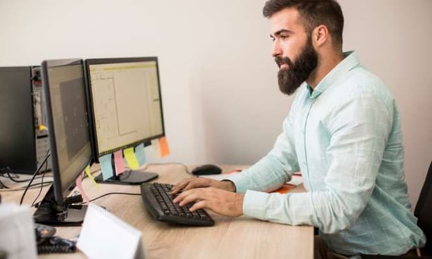 A man sitting at a desk looking at two computer monitors while resting his hands on a keyboard. The monitors have colored sticky notes on them.