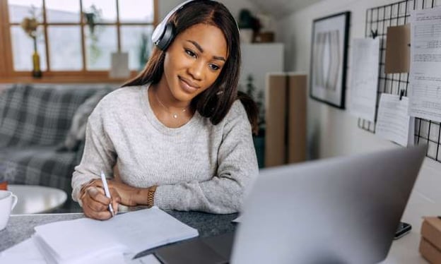 Woman wearing headphones and gray sweater is in a living room at a desk, looking at a laptop monitor and taking notes with a pen on a notepad.