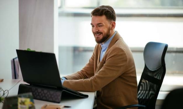 Man in a brown jacket is sitting at a desk looking at a laptop screen while smiling. He is seated in a black office chair. Window in background.