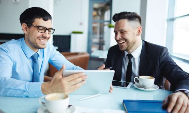 Two men looking at a tablet, smiling. They are are drinking coffee at a table.