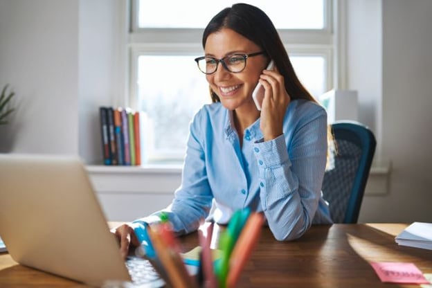 Woman wearing glasses is seated at wooden desk, looking at laptop, cell phone to her ear. She is smiling. There is a window behind her.