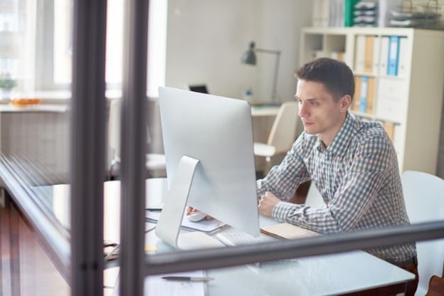 Man seated at desk, wearing checkered button up shirt, looking at computer monitor. Window and bookshelves in background.