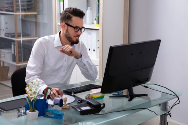 Man seated at a glass top desk, wearing glasses and white button up shirt, looking at laptop monitor. Various office items on desk.