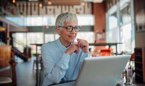 Woman seated at a table in a coffee shop, weraing glasses and blue button up shirt, looking at a laptop screen. She has a glass of water next to her.