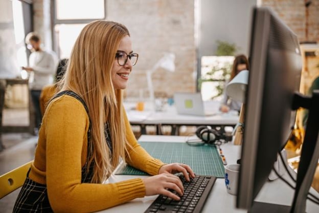 Woman wearing glasses and yellow top, seated at desk, typing on a computer keyboard. Other people and windows are in the background.