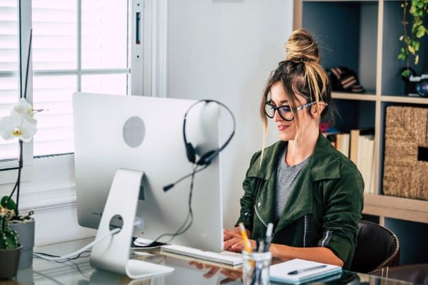 Woman wearing glasses with hair in a bun, typing on computer keyboard. A pair of headphones with a mic hangs on the side of the computer monitor.