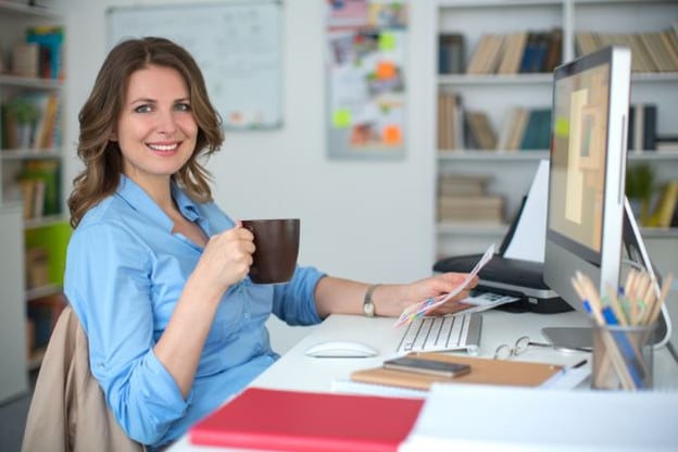 Woman seated at desk, in front of computer, holding papers in one hand and a coffee cup in the other. She is wearing a blue button up shirt.