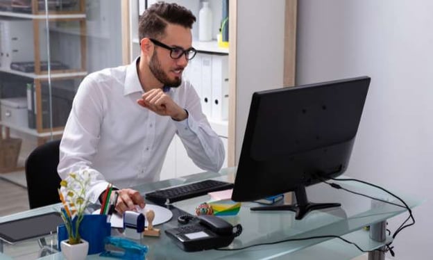 Man wearing glasses, seated at a desk, looking at a computer monitor. There are bookshelves in the background behind him.