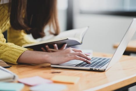Woman is leaning over onto desk, one hand on laptop, the other holding an open book. She is wearing a yellow sweater. PostIt notes on the table.
