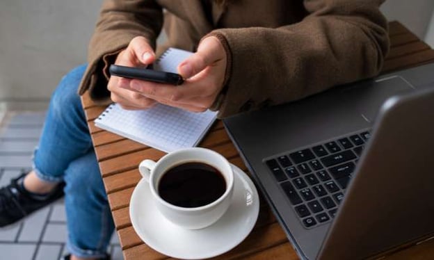 A woman sitting at a table with a laptop, a black coffee and a notebook on the table. She is engaged with her mobile device in her hands. 