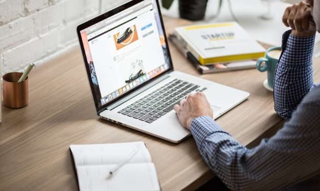 A man sits at a desk working on a laptop with text and images on the screen. On the desk are also a notebook, pen, books, and a coffee cup and saucer.