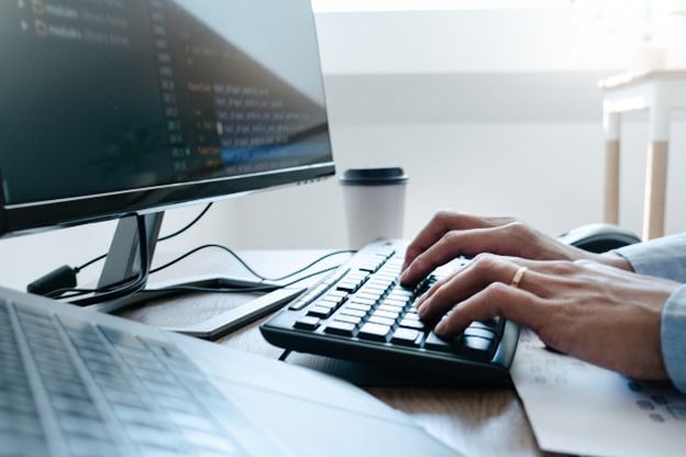 Hands typing on keyboard, there is a wedding ring on the left hand, there is a coffee cup in the background on the desk.