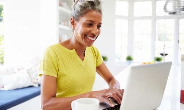 Woman seated at desk, typing on laptop, smiling, windows in the background, and a coffee cup in the foreground.