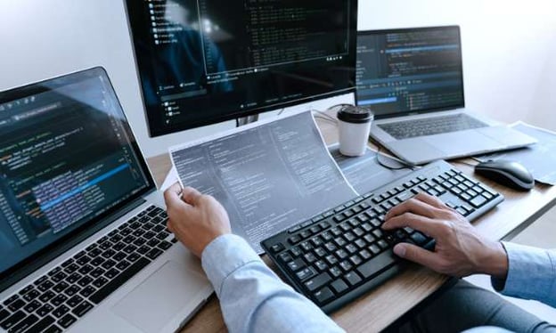 Man working at a desk on two laptops and a desktop while one hand is on a keyboard and the other hand is holding a piece of paper. 