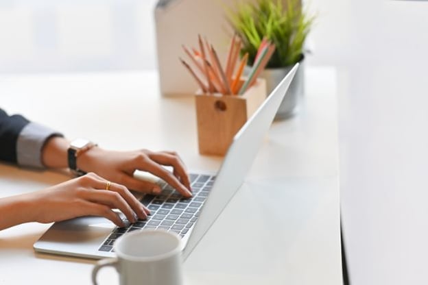 Hands typing on a laptop, there is a coffee mug in the foreground, and a pencil holder with pencils and a potted plant in the background.