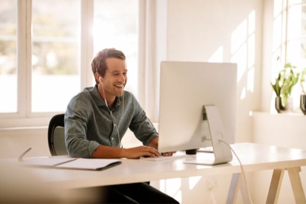 Man wearing earphones is seated at desk is smiling at his computer monitor. There is a notepad on the desk. Windows and a potted plant in background.