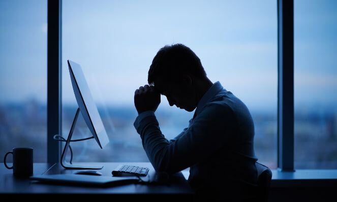 Silhouette of a person looking sad while seated at their desk in front of their computer monitor.