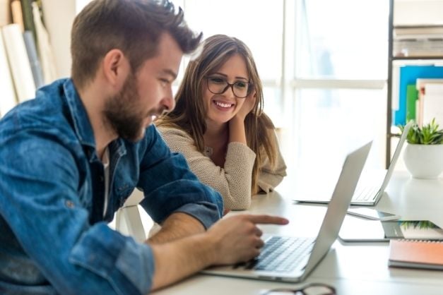 Man and woman seated a desk near window; man is pointing at laptop screen and smiling; woman is doing the same.