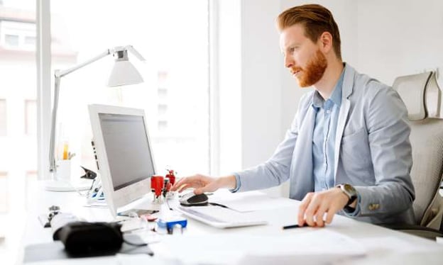 A man with red hair and a beard is seated at a desk and working on a computer. One hand is on the mouse, the other on a pen. A window is next to him.