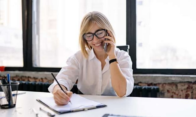 A woman sits at a table wearing glasses while talking on a cell phone and writing notes on a clipboard with paper. 