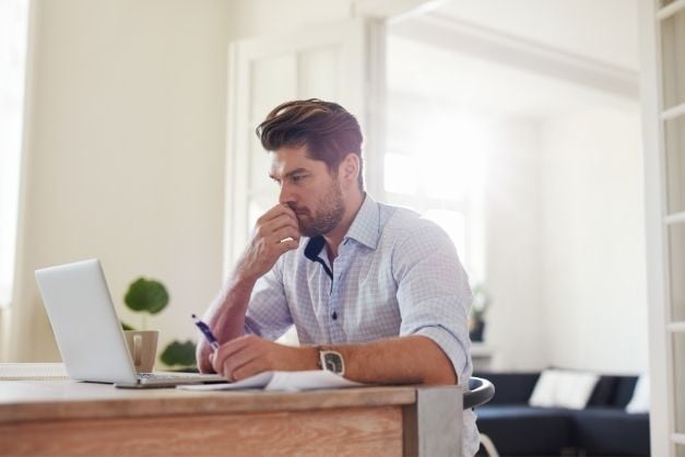 Man seated at desk, thinking while looking at laptop screen and holding a pen.