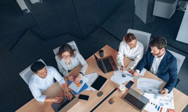 A bird's eye view of an office table with four people working. Two people are on one side of the desk, and the other two people are on the other.