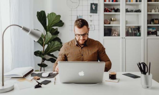 A man is sitting at a desk working on a laptop. On the desk are a book, a cup of coffee, a large lamp, and a container of pens.